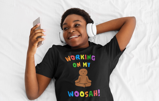 Young woman laying on a bed listening to music via headphones wearing a Working On My Woosah t-shirt
